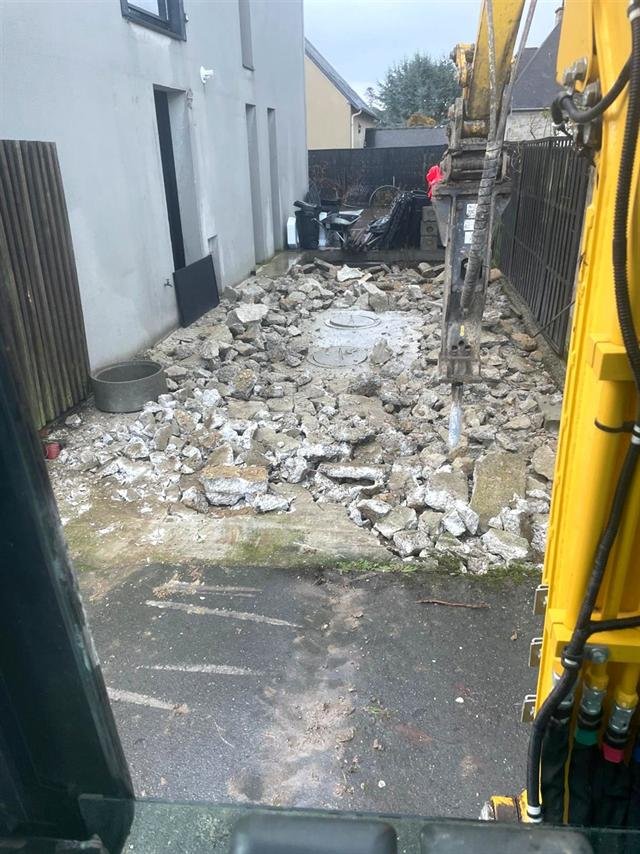 Brise-roche hydraulique cassant une dalle béton le long d'une maison, vu depuis la cabine